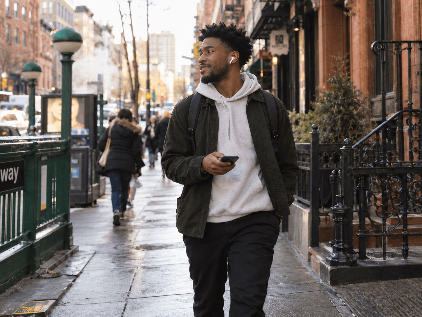 Smiling man with an earbud walking down a city sidewalk with his phone in hand